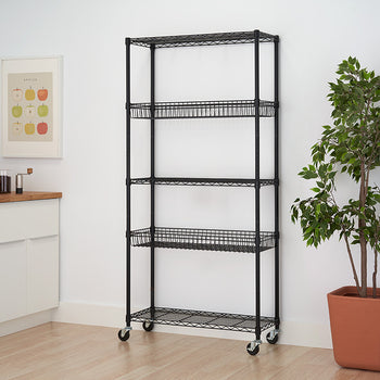 A black metal wire shelving unit with four tiers, two of which are wire baskets, stands against a white wall. The unit is on wheels and is positioned near a kitchen counter and a potted plant.