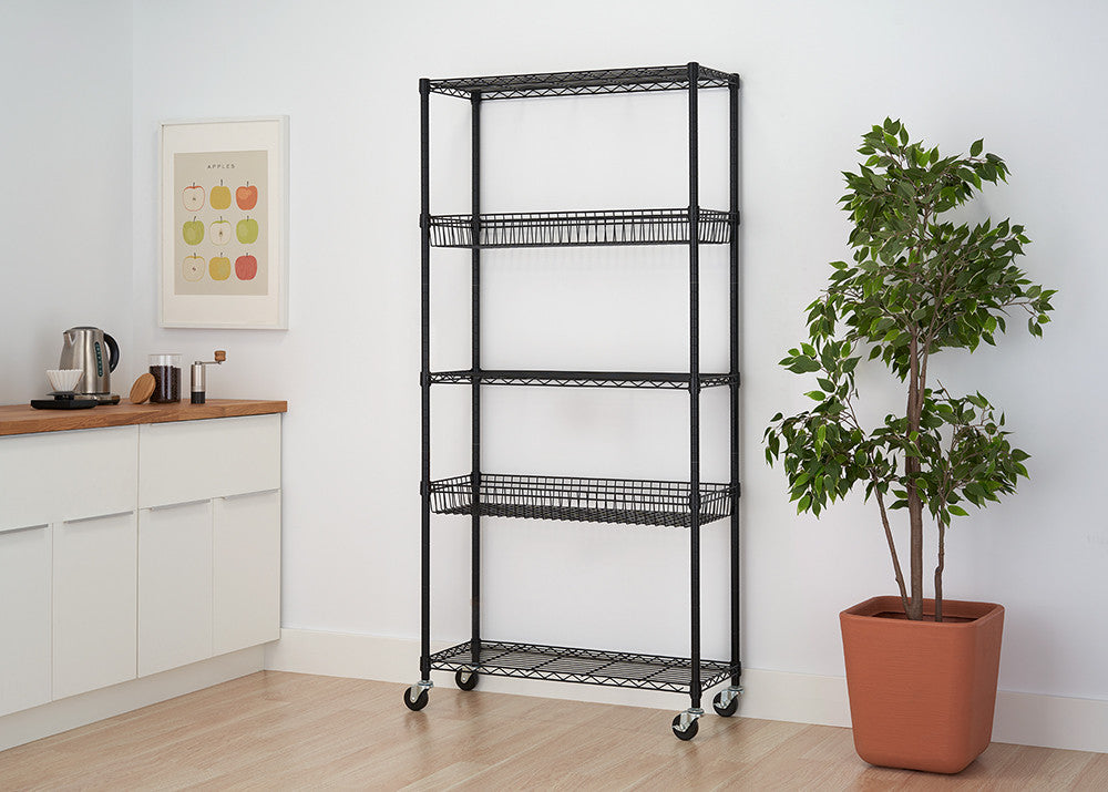A black metal wire shelving unit with four tiers, two of which are wire baskets, stands against a white wall. The unit is on wheels and is positioned near a kitchen counter and a potted plant.