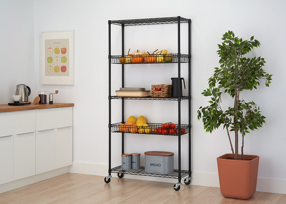 A black wire shelving unit with five tiers is shown against a white wall in a kitchen setting. The shelves hold various items including fruits, plates, and canisters, and the unit is on wheels.