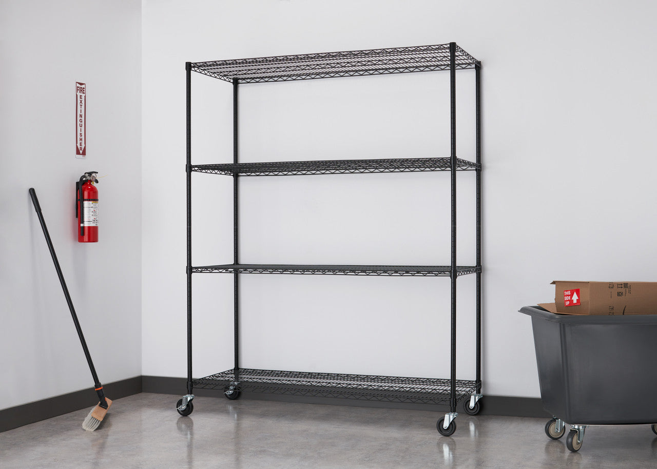 A black wire shelving unit with four tiers and wheels stands against a white wall in a room with a gray floor. A red fire extinguisher and a broom are visible to the left, and a gray bin with a cardboard box is on the right.