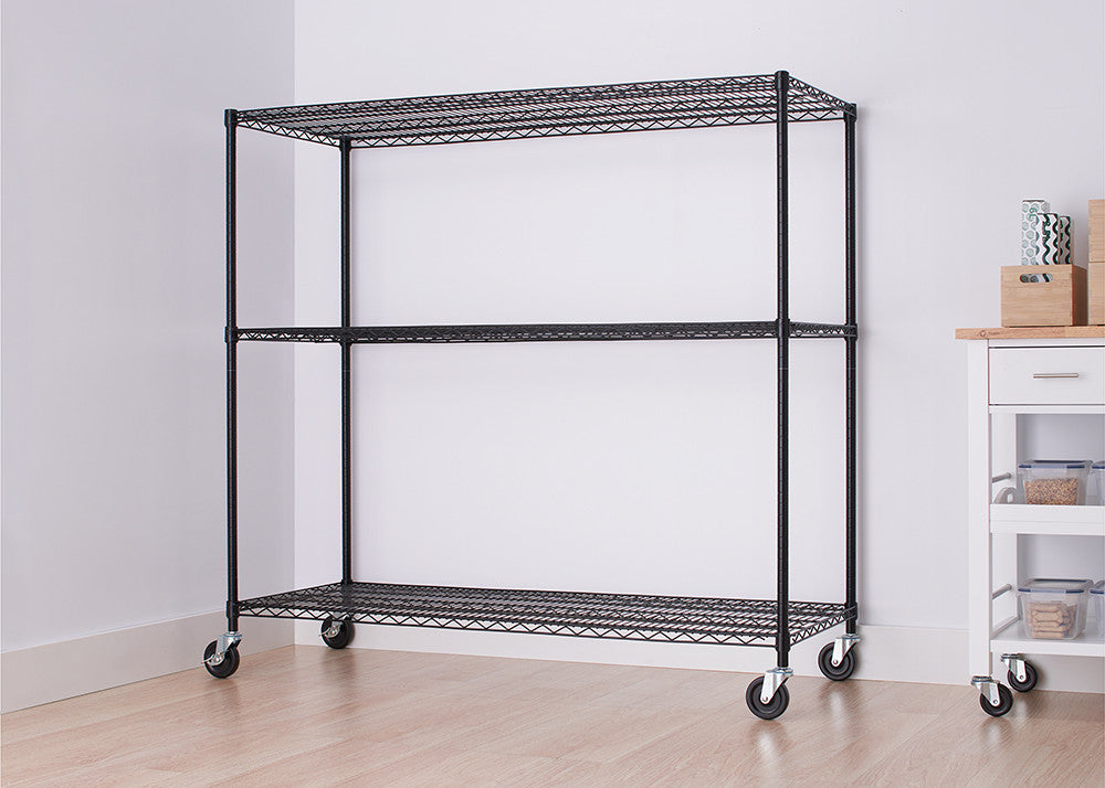 A black wire shelving unit with three tiers and wheels is centered against a white wall. The unit is empty, and a partial view of a white cabinet with drawers is visible on the right side of the image.