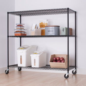 A black wire shelving unit with three tiers is shown in a room, holding various kitchen items. The shelving unit has wheels and is positioned against a white wall next to a white cabinet with a wooden top.
