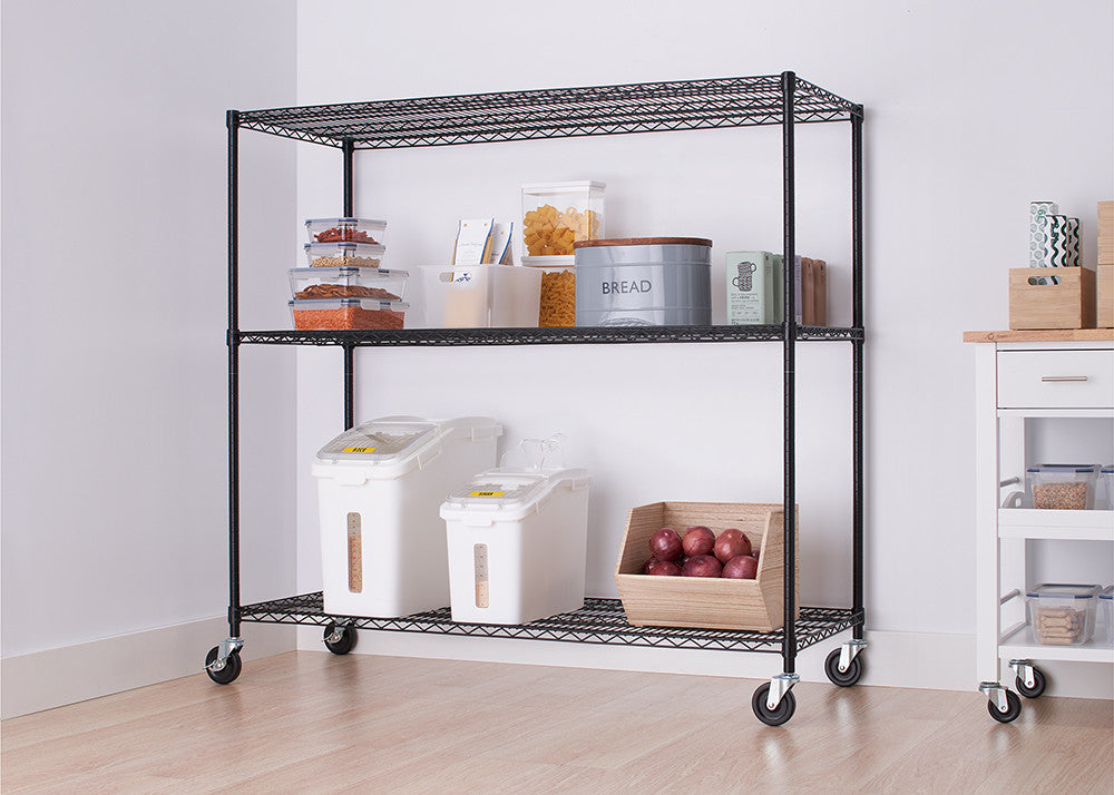 A black wire shelving unit with three tiers is shown in a room, holding various kitchen items. The shelving unit has wheels and is positioned against a white wall next to a white cabinet with a wooden top.