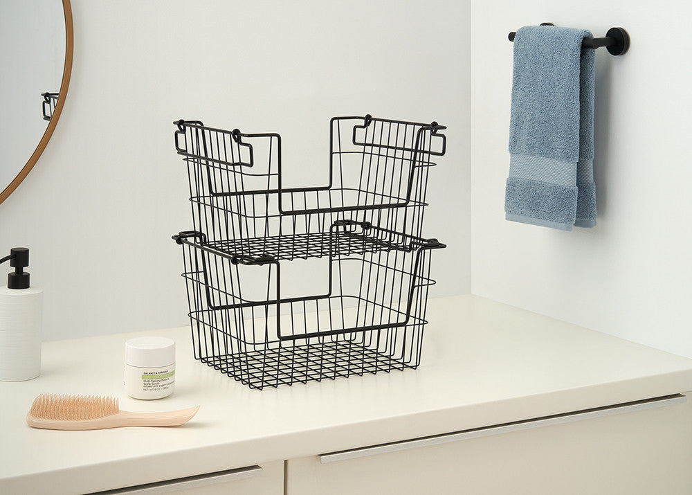 A bathroom counter features two black wire baskets stacked on top of each other. A light blue towel hangs on a rod to the right, and a round mirror is partially visible on the left.