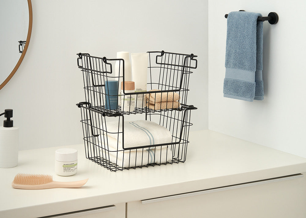 A bathroom counter features two black wire storage baskets stacked on top of each other, holding towels and toiletries. A light blue towel hangs on a rod in the background.