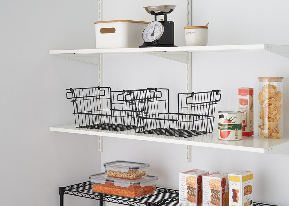 Two black wire baskets are arranged side-by-side on a white shelf against a white wall, with other kitchen items visible on the shelves above and below. The baskets are open and empty, showcasing their wire construction and stackable design.