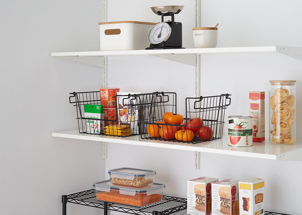Two black wire baskets sit on a white shelf, each holding various food items like oranges, canned goods, and packaged snacks. A white scale and other containers are visible on the shelf above. The image shows a well-organized pantry with a focus on storage solutions.