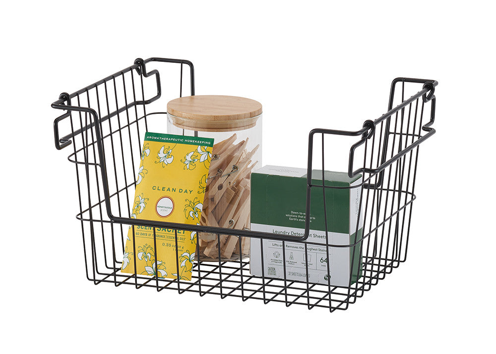 A black wire basket with a square base and open sides sits on a white background, containing a yellow packet, a glass jar with wooden clothespins, and a green box. The basket has two handles that extend upwards and slightly outwards, designed for stacking.