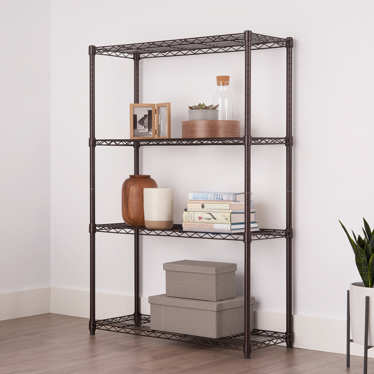 A four-tier dark bronze wire shelving unit is shown in a room with white walls and a light wood floor. The shelves hold various decorative items, including books, vases, boxes, and a framed picture.