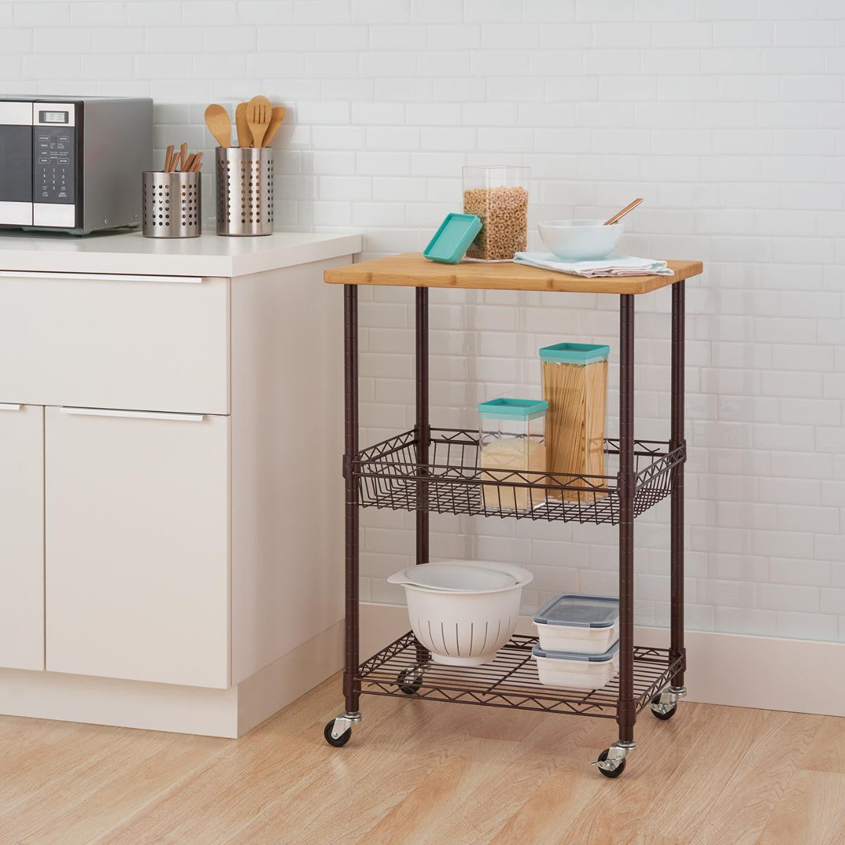 A three-tier dark bronze kitchen cart with a wooden top and a wire basket is positioned in a kitchen setting. The cart is equipped with wheels for mobility and holds various kitchen items.