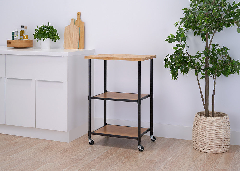 A modern kitchen setting with a rolling kitchen cart featuring a wooden top, two wooden shelves, and a black metal frame. The cart is positioned between a white kitchen counter with herbs and cutting boards and a potted green plant in a woven basket.