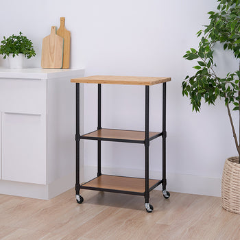 A modern kitchen setting with a rolling kitchen cart featuring a wooden top, two wooden shelves, and a black metal frame. The cart is positioned between a white kitchen counter with herbs and cutting boards and a potted green plant in a woven basket.