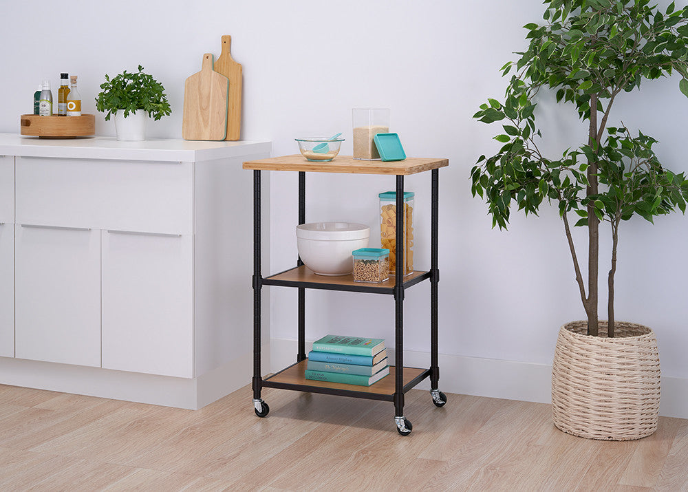A modern kitchen features a rolling cart with a wooden top and black metal frame, holding bowls, cereal containers, and books. The space is decorated with a potted plant, white cabinets, and wooden cutting boards.