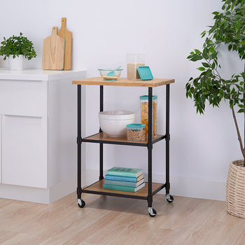 A modern kitchen features a rolling cart with a wooden top and black metal frame, holding bowls, cereal containers, and books. The space is decorated with a potted plant, white cabinets, and wooden cutting boards.