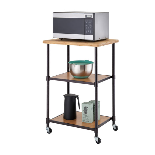 A three-tier kitchen storage cart with a wooden top, black metal frame, and caster wheels holds a microwave on the top shelf. The middle and bottom shelves store a mixing bowl, a black pitcher, and two green boxes.