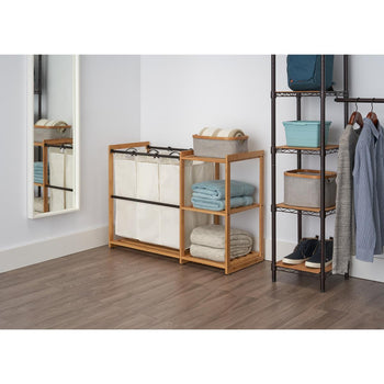 A Trinity Bamboo Laundry Station stands against a white wall, featuring three canvas hamper bags on the left and two shelves with folded towels and blankets on the right. To its right, a dark metal shelving unit holds various items, including a blue container, a gray bin, and two pairs of shoes, with clothes hanging from an attached rail.