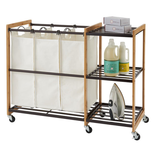 A Trinity 3-Bag Bamboo Laundry Station with Wheels. It features three removable laundry bags on the left and a shelving unit on the right holding laundry detergent, an iron, and folded boxes of dryer sheets.