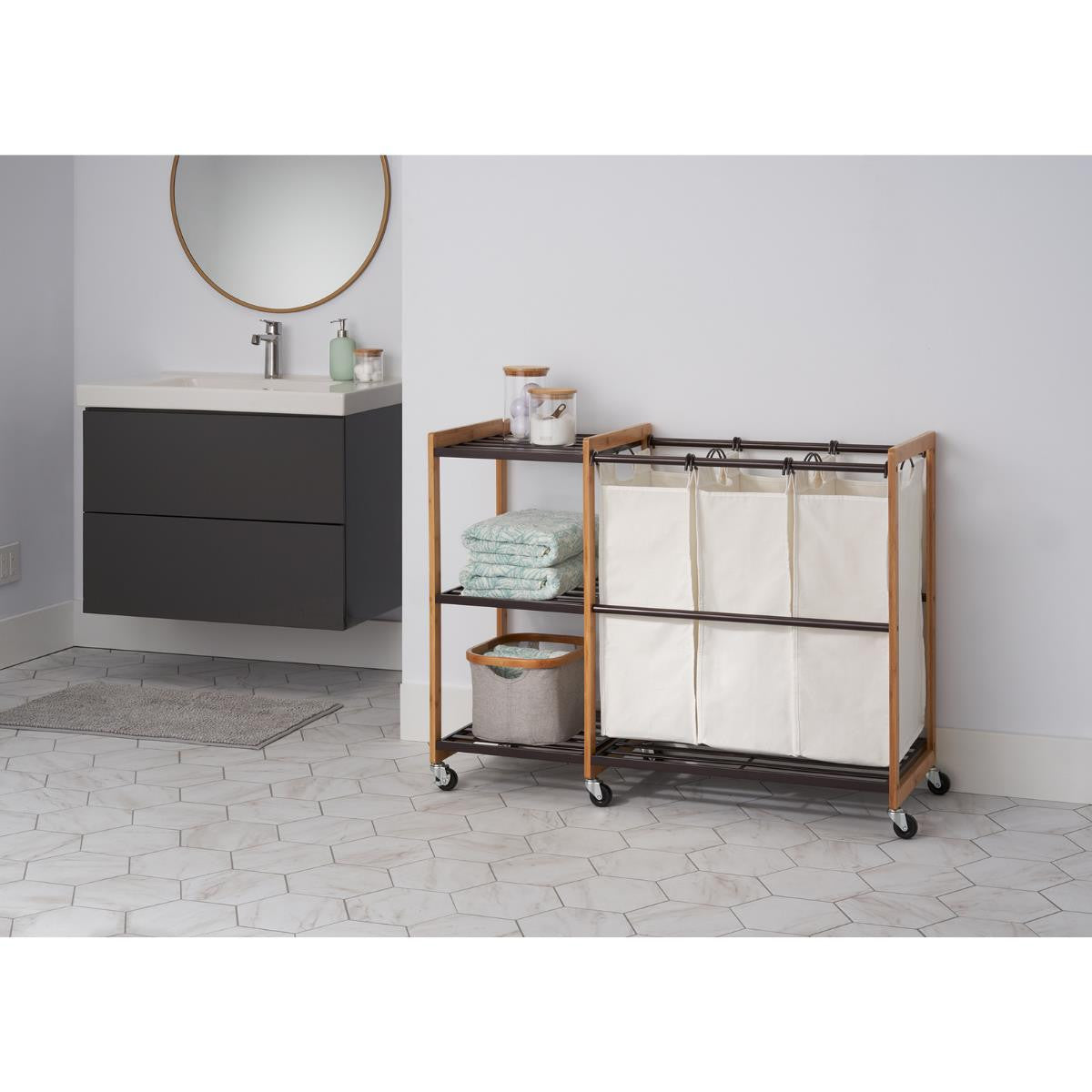A Trinity 3-Bag Bamboo Laundry Station with Wheels, featuring three white laundry bags, sits next to a matching two-shelf unit. Both are made with bamboo frames and dark metal accents, positioned on a light hexagonal tiled floor in a bathroom with a floating dark wood vanity.