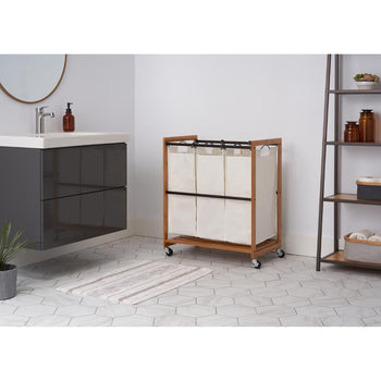 A natural bamboo Trinity 3-Bag Laundry Cart with cream-colored laundry bags and a black metal frame is positioned on hexagonal white bathroom tiles. In the background, a modern floating vanity with a white sink and dark gray drawers is visible to the left, and a multi-shelf unit with various brown jars and bottles is on the right.