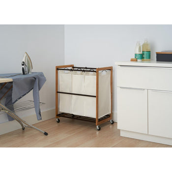 A 3-bag laundry sorter with a wooden frame and cream-colored bags stands on wheels in a laundry room. An ironing board with an iron and a blue and white striped shirt is visible on the left, and a white cabinet with bottles of laundry detergent sits on the right.