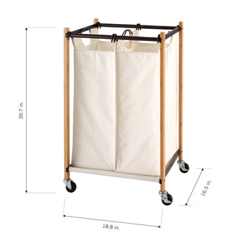 A beige, two-compartment laundry hamper with a light wooden frame and black metal accents is shown from an eye-level perspective. The hamper has black wheels and its dimensions are labeled as 30.7 inches in height, 18.8 inches in width, and 16.5 inches in depth.