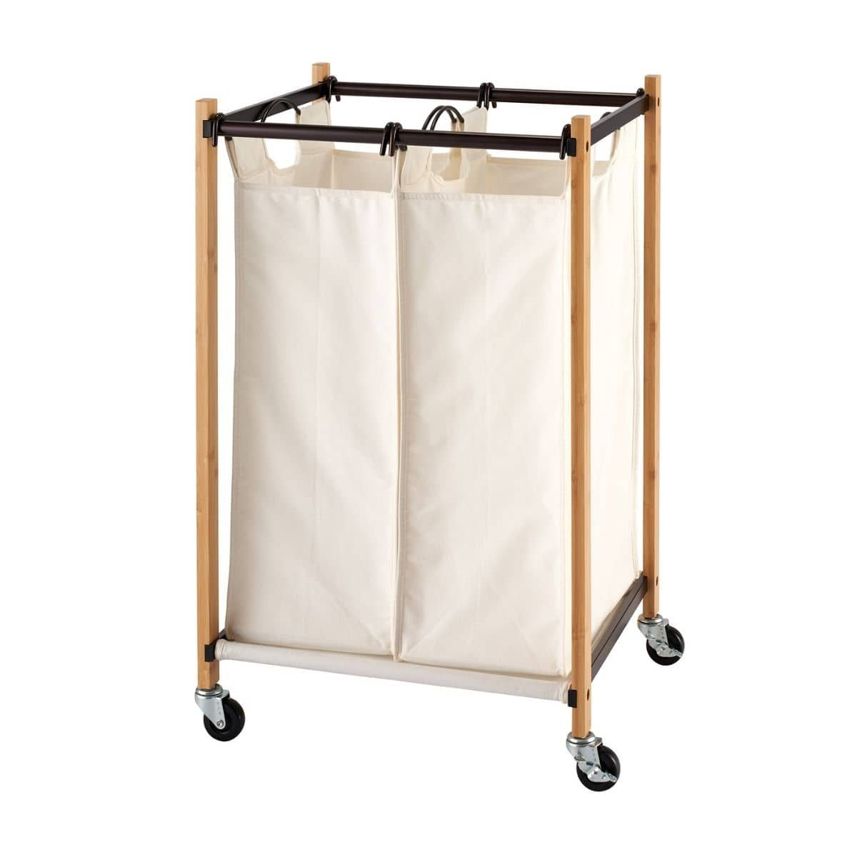 A laundry cart with two cream-colored fabric bags hangs from a dark brown metal frame supported by light brown bamboo poles. The cart has four black wheels for mobility.