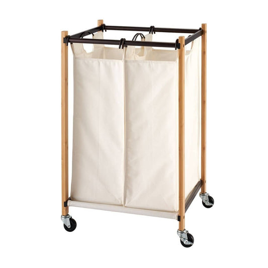 A laundry cart with two cream-colored fabric bags hangs from a dark brown metal frame supported by light brown bamboo poles. The cart has four black wheels for mobility.