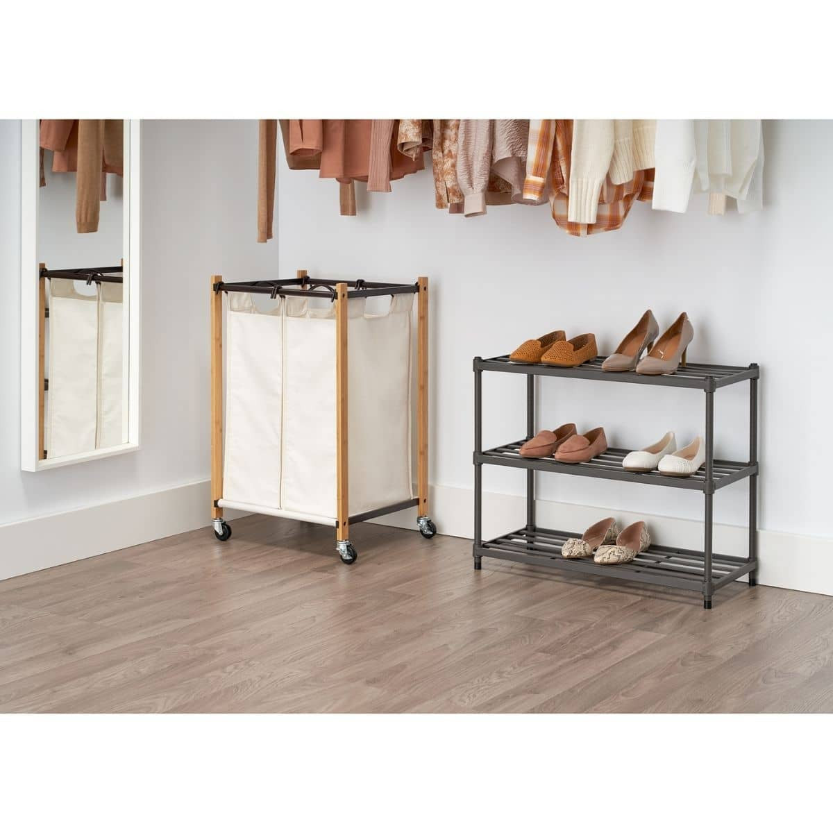 A low-angle, indoor shot shows a gray, three-tiered metal slat shoe rack against a white corner wall, holding nine pairs of shoes on its shelves. To the left, a tall, rectangular laundry hamper with a light canvas bag and wooden frame on wheels sits on a light brown wood-look floor, near a white-framed mirror and hanging clothes in the background.