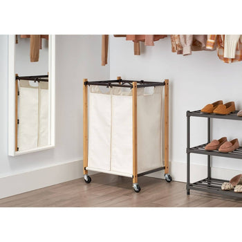 A light-colored, double-compartment laundry hamper with a bamboo frame and white canvas bags sits on a light wood floor. The hamper has black trim and rolling casters for mobility.