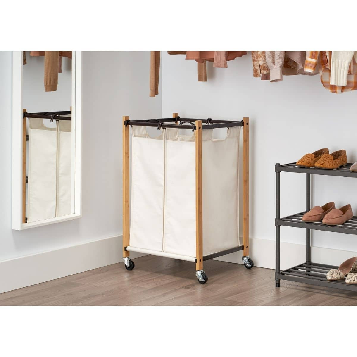 A light-colored, double-compartment laundry hamper with a bamboo frame and white canvas bags sits on a light wood floor. The hamper has black trim and rolling casters for mobility.