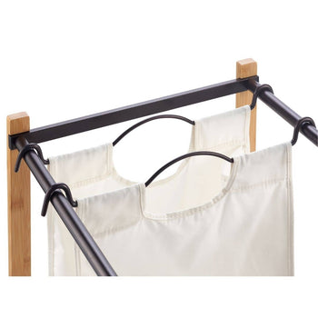 A close-up shows a double laundry hamper with two off-white canvas bags hanging from black metal frames and hooks attached to light brown wooden supports. The focus is on the top portion of the hamper, highlighting the construction and materials.