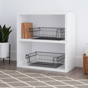 A white, two-tiered cube shelf unit holds two black wire baskets on its shelves. A potted plant and a woven basket are visible on the periphery.