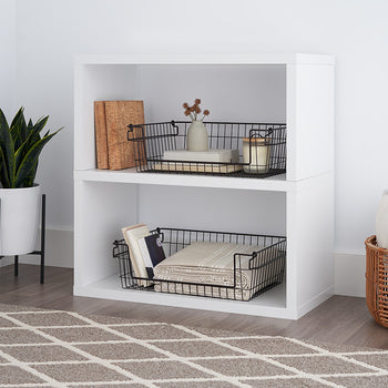 A bright, modern room features a white cube storage unit with two dark bronze wire baskets inside, each holding various items. A potted plant stands to the left, and a woven basket sits to the right, all on a patterned rug.