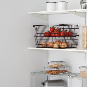A close-up shot of a white wall with two white shelves. The shelves hold black wire baskets filled with tomatoes and onions, as well as clear containers with various dry goods.