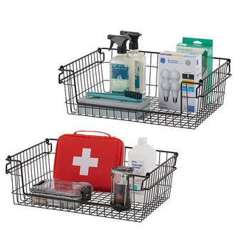 Two dark bronze wire baskets are stacked on a white background, each filled with various household items. The top basket contains cleaning supplies and light bulbs, while the bottom basket holds a first aid kit and other miscellaneous items.