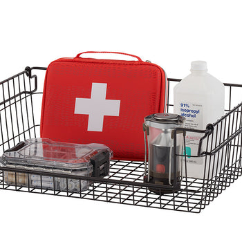 A black wire basket on a white background, filled with various items including a red first aid kit, a clear plastic container, a metal thermos, and a white bottle labeled 