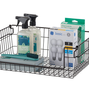 A black wire basket on a white background, filled with cleaning supplies, light bulbs, and a green toothbrush. The basket has a simple, open design with a flat base and slightly flared sides.
