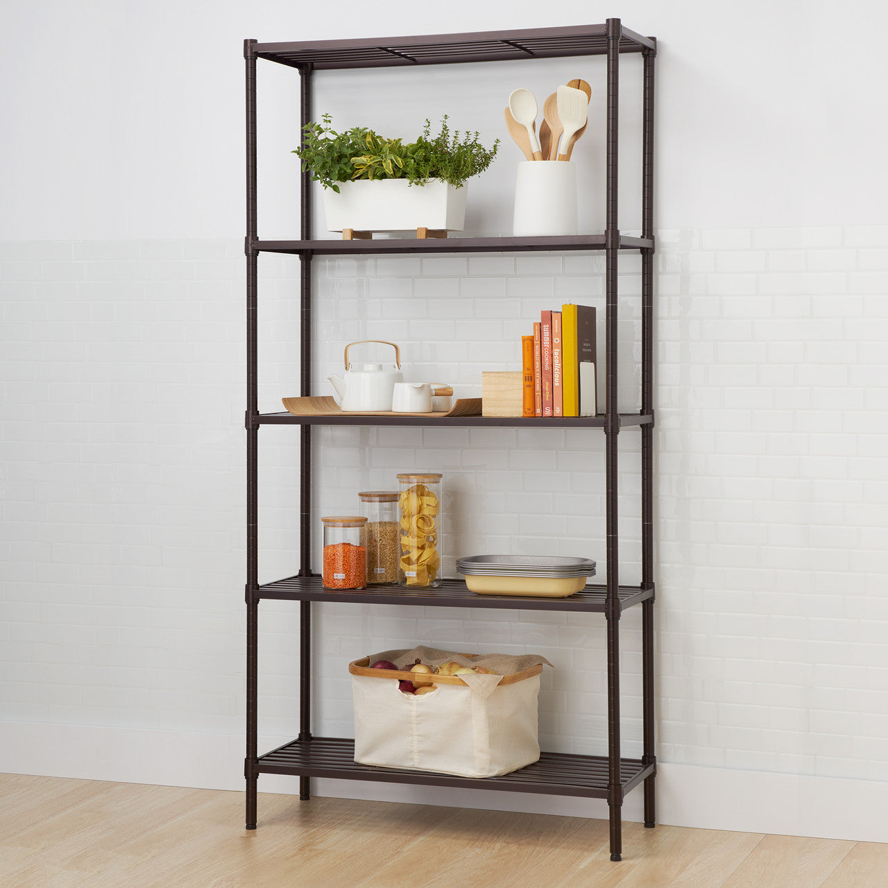 A five-tier dark bronze metal shelving unit stands against a white subway tile wall. The shelves are decorated with kitchen and home items, including a plant, books, dishes, food containers, and a fabric bin.