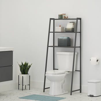 A three-quarter view of a leaning dark metal A-frame over-the-toilet space saver in a contemporary bathroom with a dark grey floating vanity and geometric floor tiles. The space saver features three wood-toned shelves with various toiletries, including storage bins, bottles, and stacked towels, and is positioned over a white toilet.