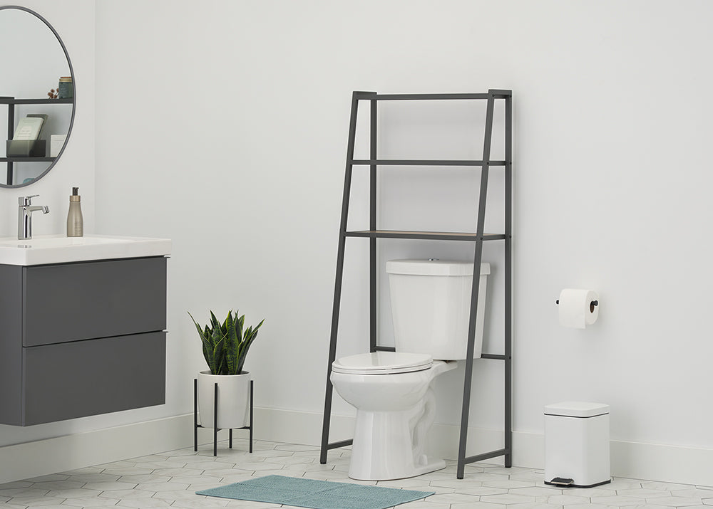 A leaning dark gray metal A-frame over-the-toilet space saver is positioned over a white toilet in a modern bathroom with grey floating cabinetry and hexagon tile flooring. The space saver has three wood-toned shelves which are empty in this version, and the rest of the scene, including a snake plant, trash can, and vanity, is visible.