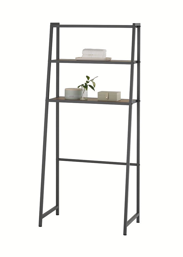 A leaning dark gray metal and light wood over-the-toilet space saver is shown against a plain white background. The bottom shelf holds folded white towels in a tray, while the top shelf displays bathroom accessories including a lidded container, a sprig of greenery in a bud vase, and a soap dish.