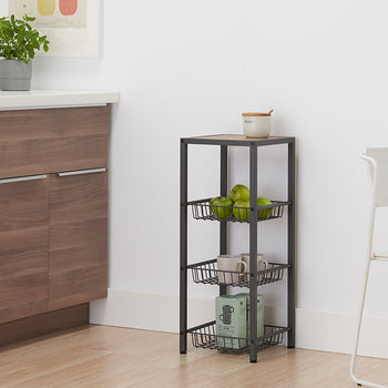 A dark gray, four-tiered metal shelving unit with wire baskets sits on a light wooden floor next to a white wall. The top shelf, made of light wood, holds a cream-colored ceramic jar, while the baskets below contain green apples, mugs, and a box.