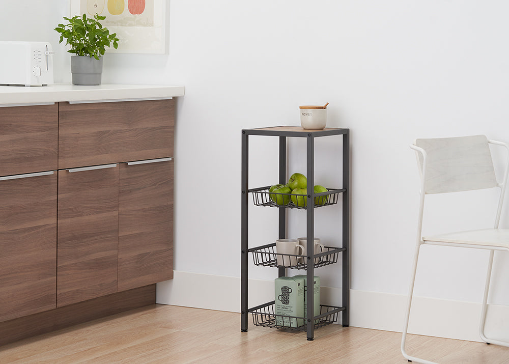 A dark gray, four-tiered metal shelving unit with wire baskets sits on a light wooden floor next to a white wall. The top shelf, made of light wood, holds a cream-colored ceramic jar, while the baskets below contain green apples, mugs, and a box.