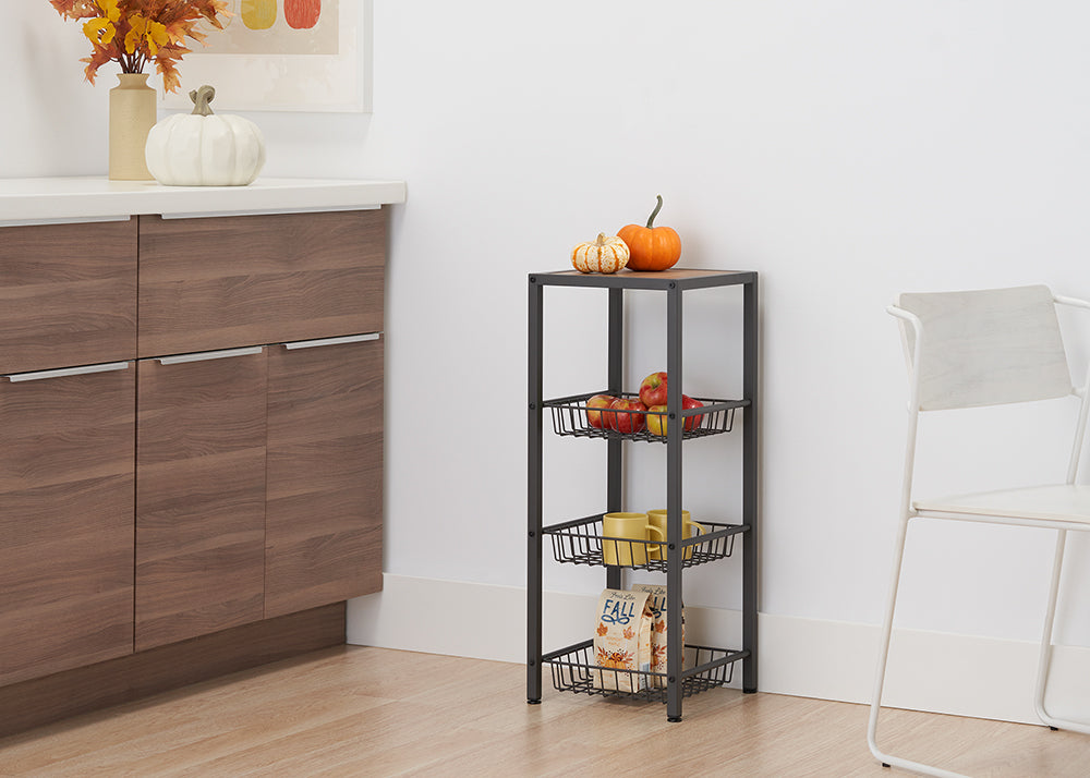 A dark gray metal shelving unit stands in a corner with a light wood top. It has three wire baskets holding apples, mugs, and a "FALL" themed bag, with two small decorative pumpkins on the top shelf.
