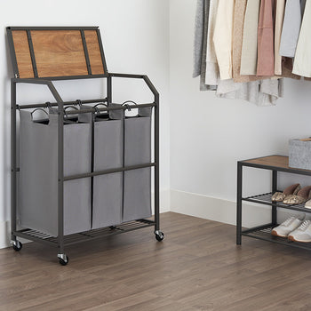 A laundry cart with three gray fabric bags and a flip-top wooden lid stands next to a closet with hanging clothes. A matching shelf with shoes and gray bins is also visible.