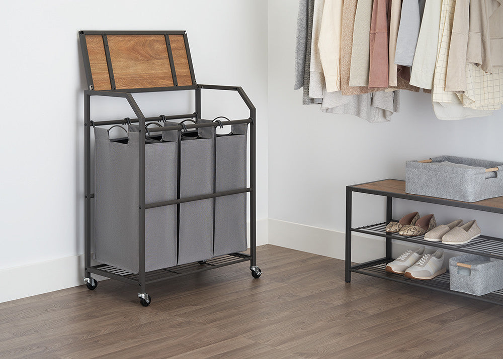 A laundry cart with three gray fabric bags and a flip-top wooden lid stands next to a closet with hanging clothes. A matching shelf with shoes and gray bins is also visible.