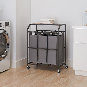 A laundry cart with three gray fabric bins is positioned in a laundry room with a washing machine and white cabinets. The cart has a dark metal frame, a wooden top, and is on wheels.