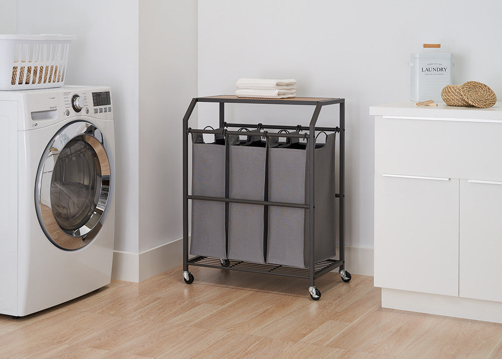 A laundry cart with three gray fabric bins is positioned in a laundry room with a washing machine and white cabinets. The cart has a dark metal frame, a wooden top, and is on wheels.