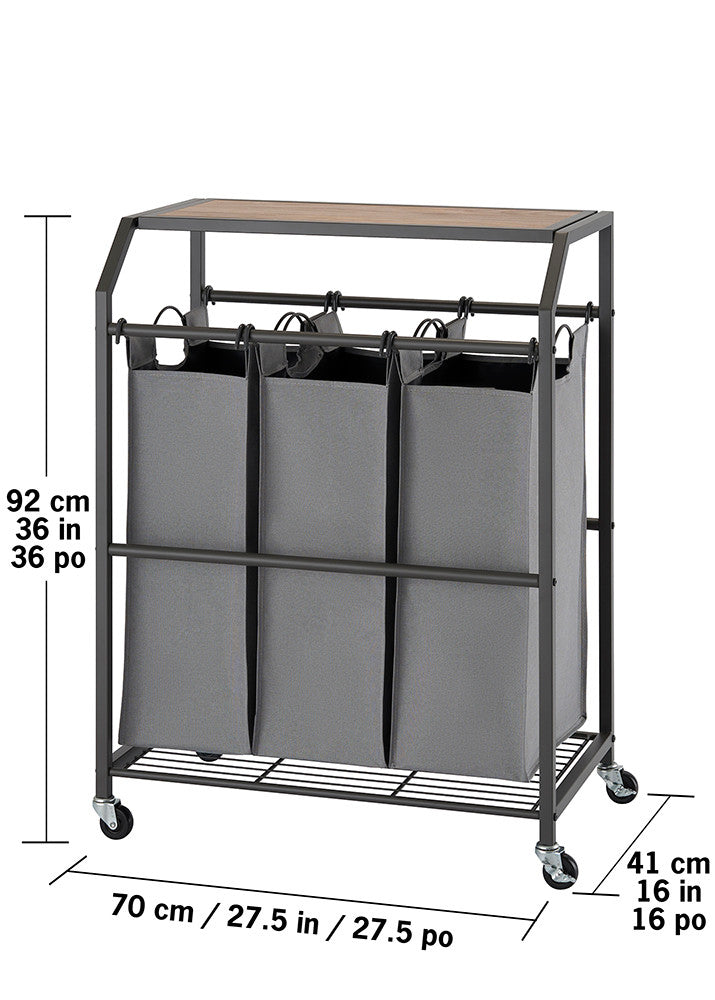 A laundry cart with three gray fabric bags hanging from a metal frame with a wooden top. The cart has wheels and measurements are shown in centimeters and inches.