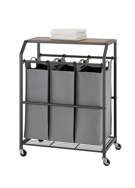 A laundry cart with three gray fabric bins hanging beneath a wooden top, all encased in a dark metal frame with wheels. A folded white towel rests on the wooden surface.